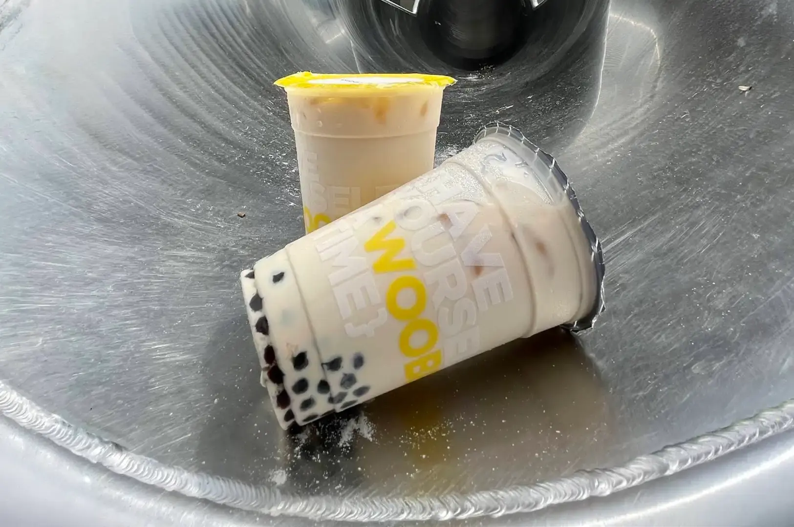 Two Woobbee bubble tea cups rest inside a stainless steel basin, with the foreground cup lying on its side to display its creamy iced milk tea and dense layer of dark tapioca pearls alongside bold yellow brand lettering that reads "HAVE YOURSELF A WOOBBEE TIME."