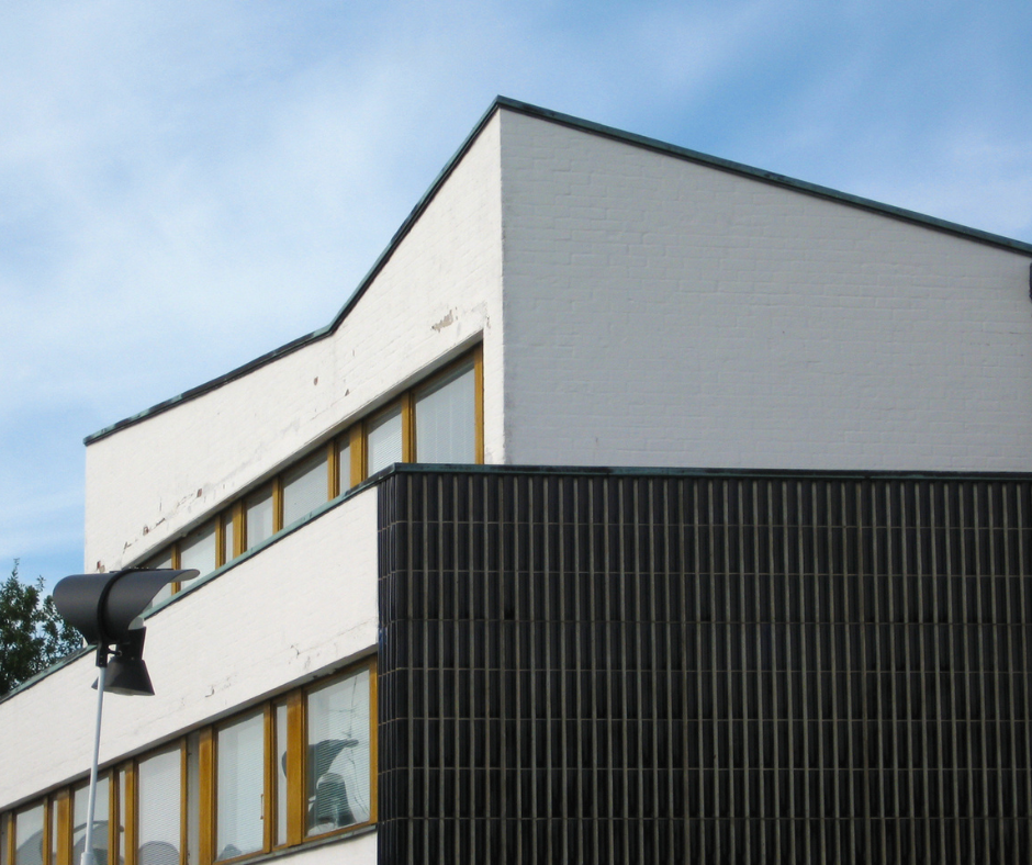Exterior view of a modernist building with white brick upper walls, long horizontal windows framed in wood, and a dark vertical-tile facade section in the foreground.