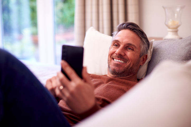 Mature man laying on a sofa and smiling at a cell phone. 