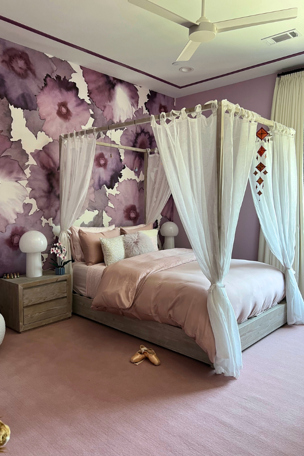 Bedroom with lavender walls and large-scale floral mural behind a canopy bed dressed in blush bedding, paired with sheer white drapery and soft pink carpet.