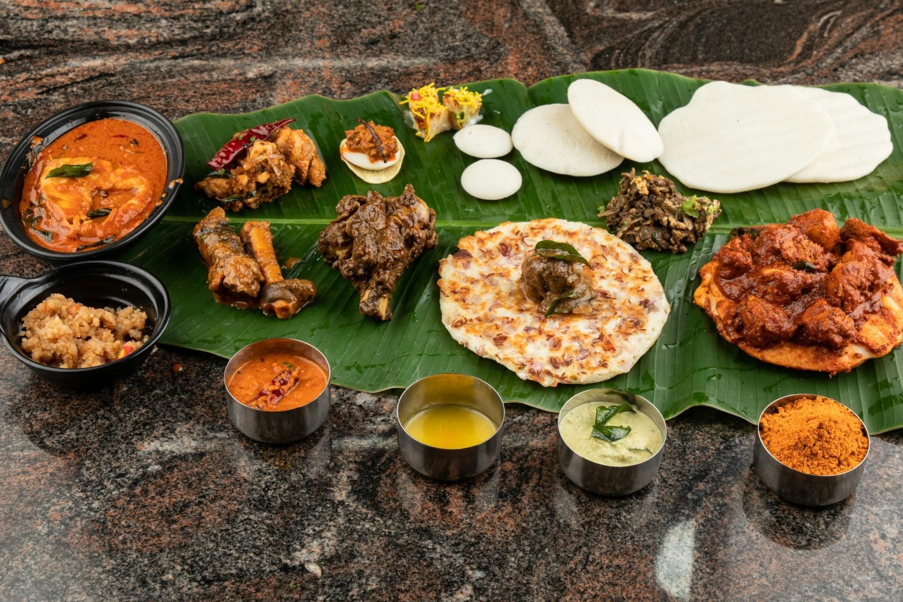  This image displays a rich assortment of South Indian dishes, including idli, uttapam, and various curries, served traditionally on a large green banana leaf. Accompanying the main items are several small metal bowls containing colorful chutneys, sambar, and spices, all set against a dark granite tabletop.