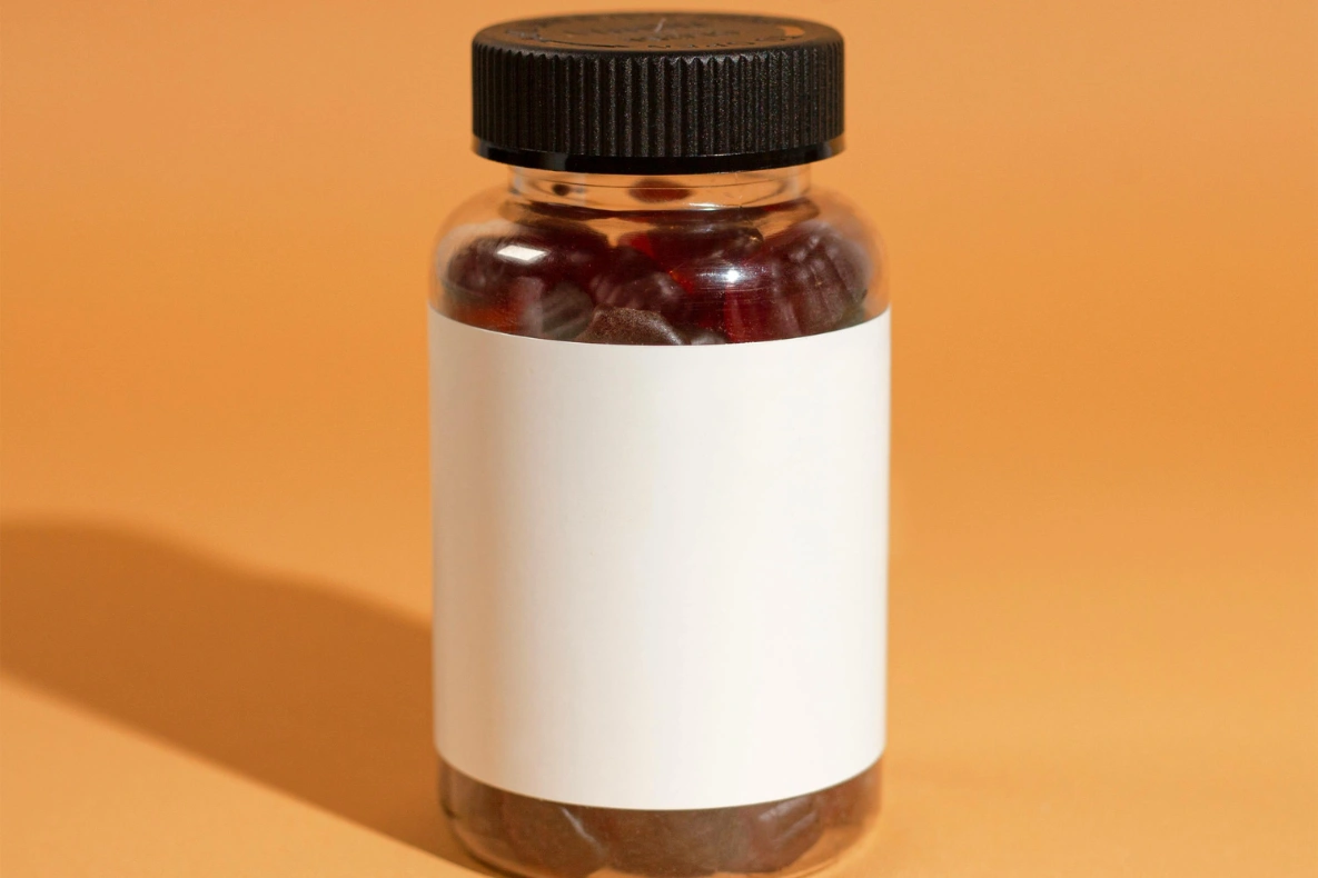A clear bottle with a black cap filled with dark red gummy supplements sits on an orange background with a shadow to the left.