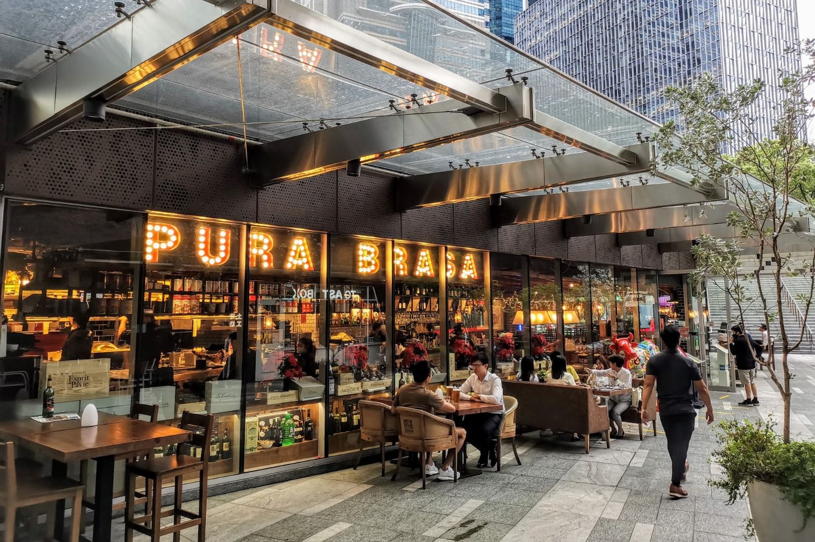 Outdoor seating of a modern restaurant with glass walls and "PURA BRASA" in bright lights. Diners enjoy meals, conveying a lively, urban atmosphere.