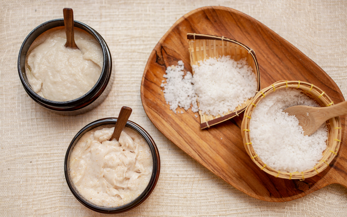 Natuurlijke scrubingrediënten op een houten dienblad, waaronder twee potten met crèmekleurige scrubs en mandjes gevuld met zeezout.