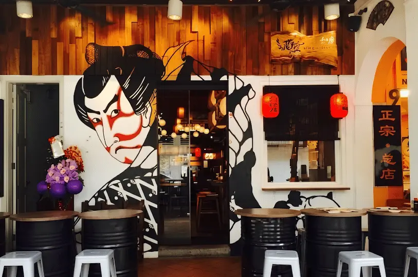 The storefront of a vibrant izakaya in Singapore, featuring a striking wall mural of a traditional Kabuki actor, warm wooden paneling, red paper lanterns, and black oil drum tables for outdoor seating.