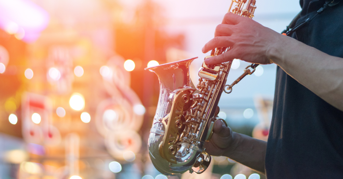 Close-up of a musician playing a saxophone at an outdoor Cape May music festival during sunset, with glowing lights and musical note decorations blurred in the background.