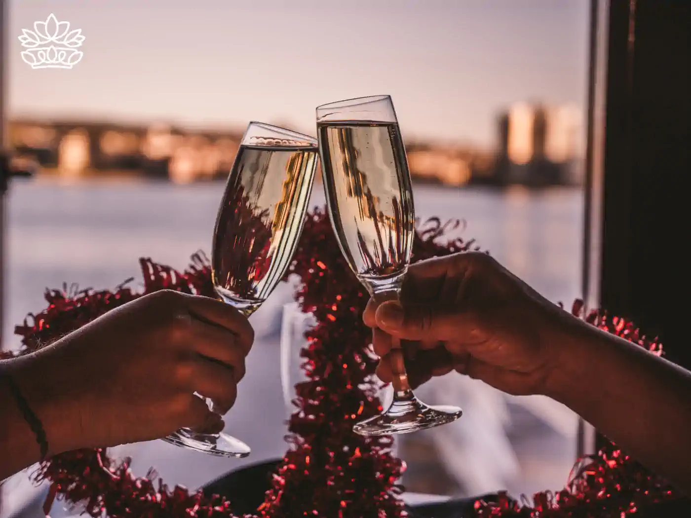 Two hands clinking champagne glasses adorned with red tinsel, celebrating Valentine's Day against a backdrop of a sunset cityscape. Fabulous Flowers and Gifts delivered with heart. Valentine's Day Flowers.