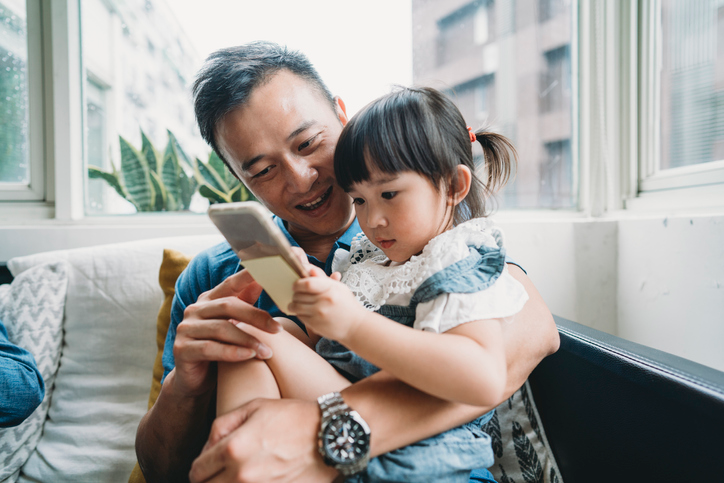 Happy young dad and daughter sitting in a white chair and looking at a cell phone. 