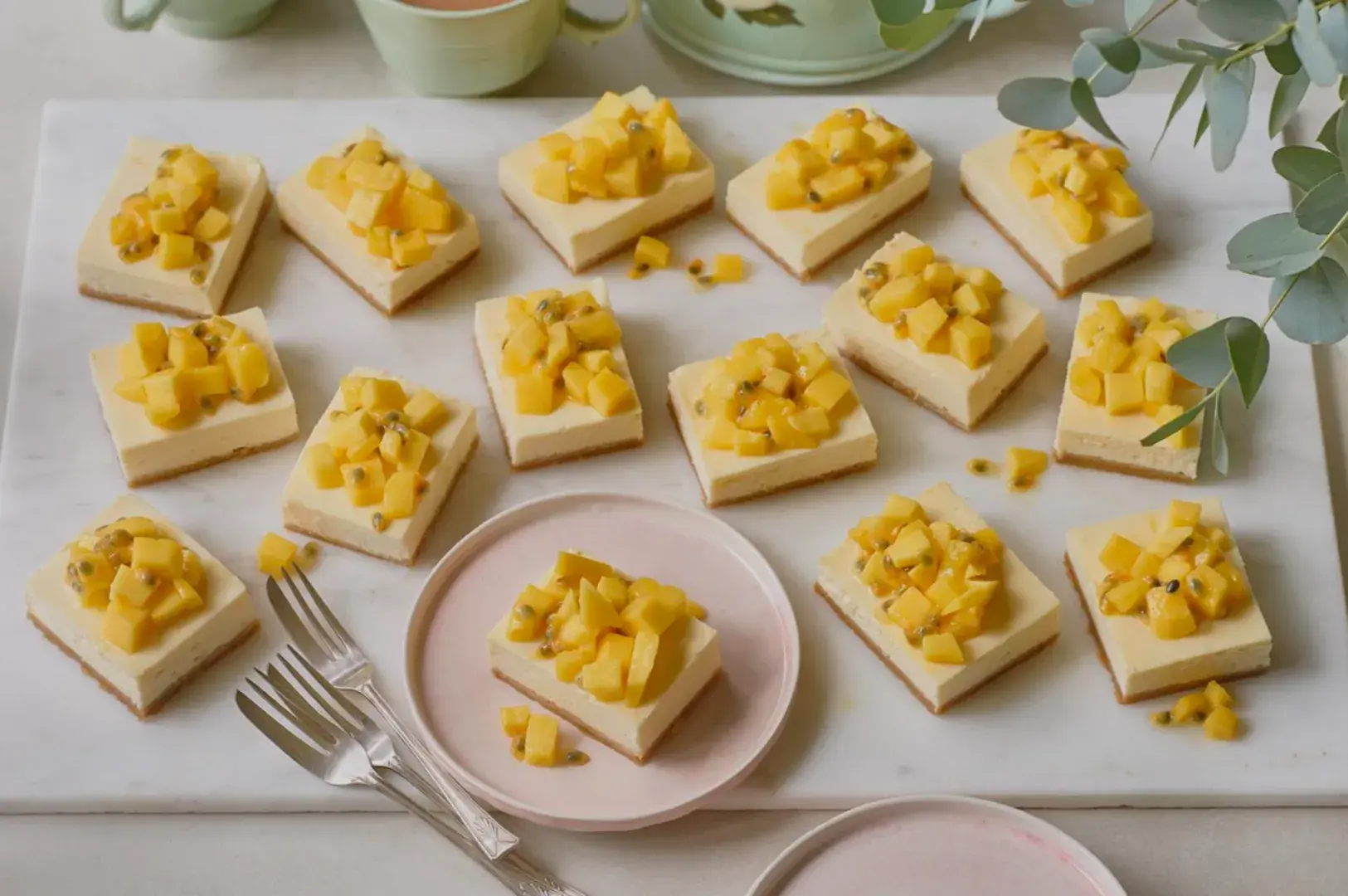 A marble platter topped with rectangular mango cheesecake squares, each adorned with diced mango pieces, with pink plates and forks on the side.