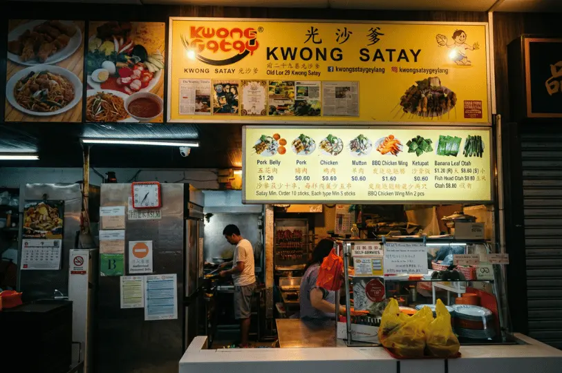 The brightly lit storefront of Kwong Satay, a hawker stall in Singapore. A large yellow signboard displays the stall's name alongside newspaper clippings and social media handles. Below it, an illuminated menu board shows pictures and prices for various skewers, including pork belly, chicken, mutton, and BBQ chicken wings. Inside the bustling stall, a cook tends to a grill in the background, while a woman at the front counter packs takeout orders into yellow plastic bags.