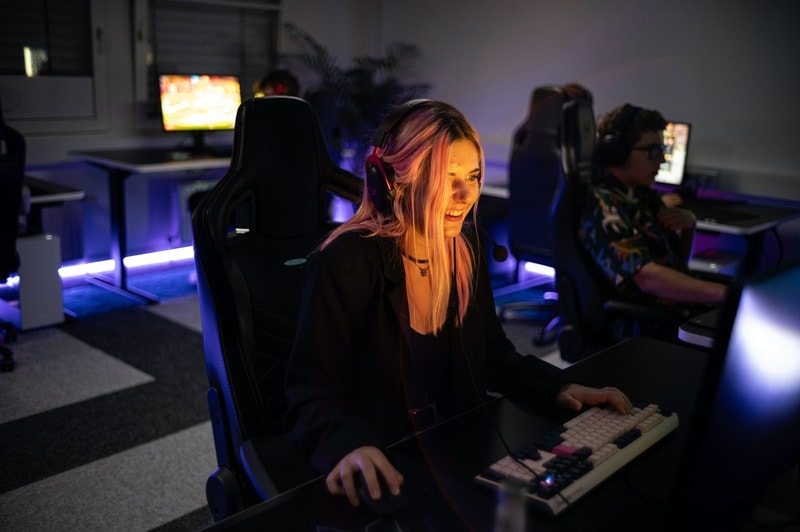 A woman with headphones smiles while playing on a computer in a dimly lit gaming room.