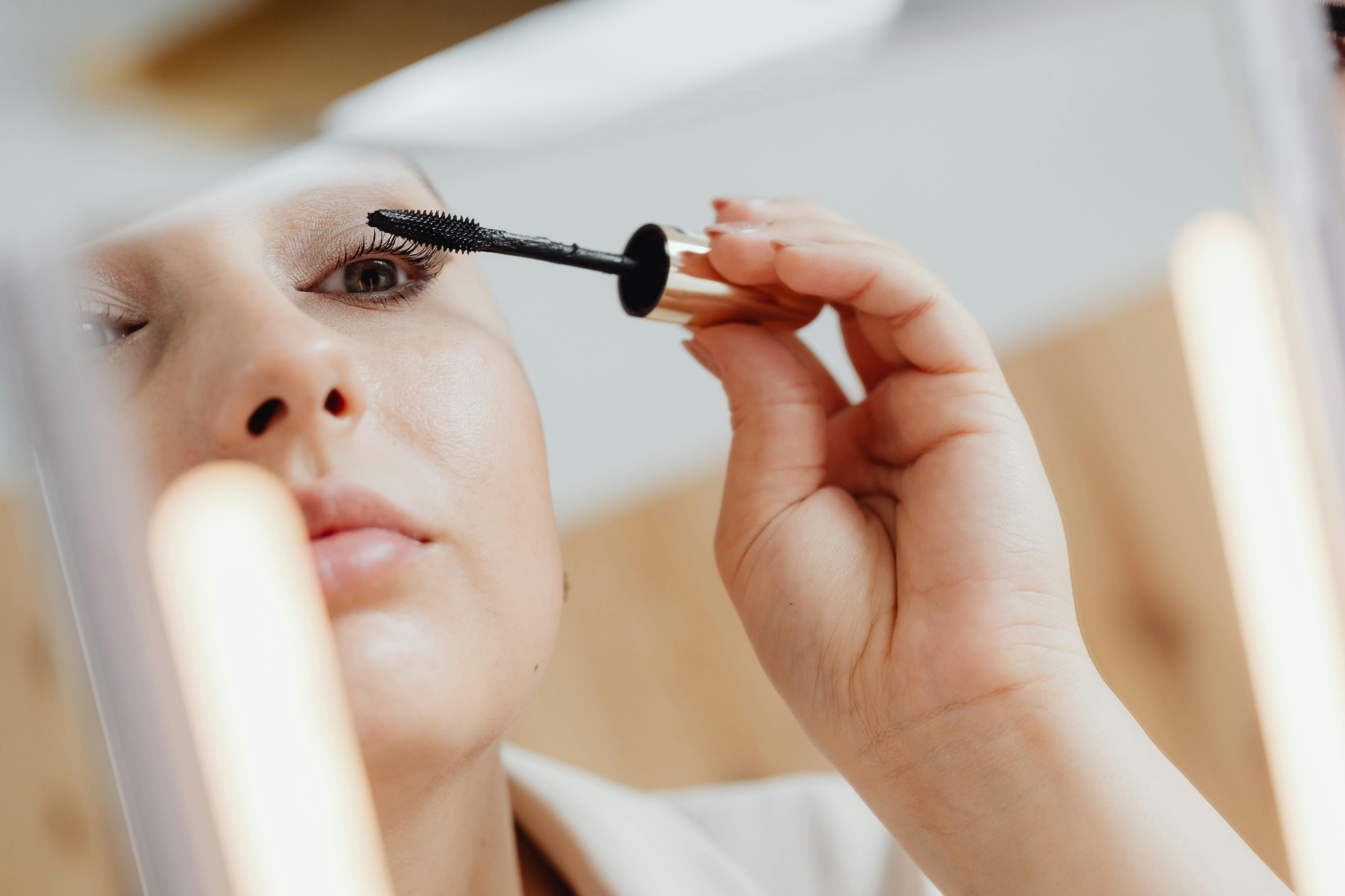A close-up, reflected image in a lighted vanity mirror showing a person applying black mascara to their upper eyelashes with a wand. The focus is on the right eye, which is open. The person's hand is holding the mascara wand near the eye. The bright lighting of the mirror illuminates the face.
