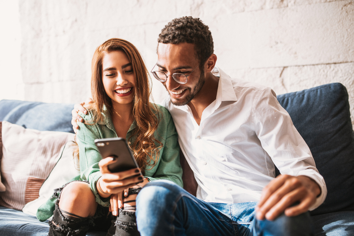 Cheerful young couple sitting on the sofa looking at a cell phone. 