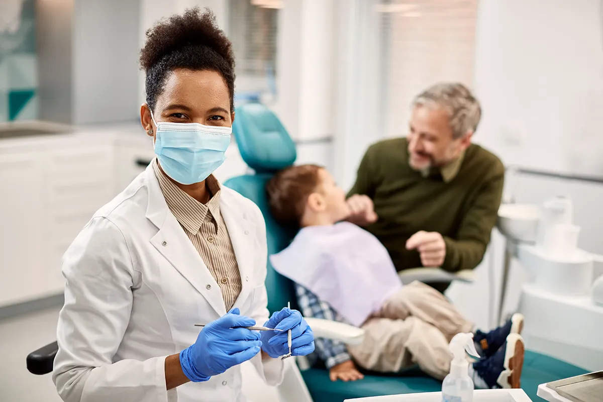 Female dental professional in foreground with dental tools while child patient receives treatment with parent support in background