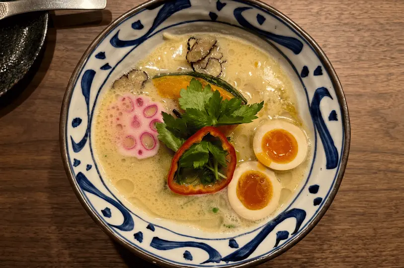 A bowl of creamy ramen with soft-boiled eggs, pink fish cake, cilantro, and sliced red pepper garnished in a blue-patterned bowl on a wooden table.