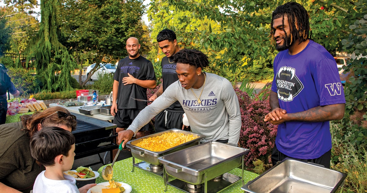 Washington Huskies football players serving food to community members at a charity event, highlighting NIL community involvement.