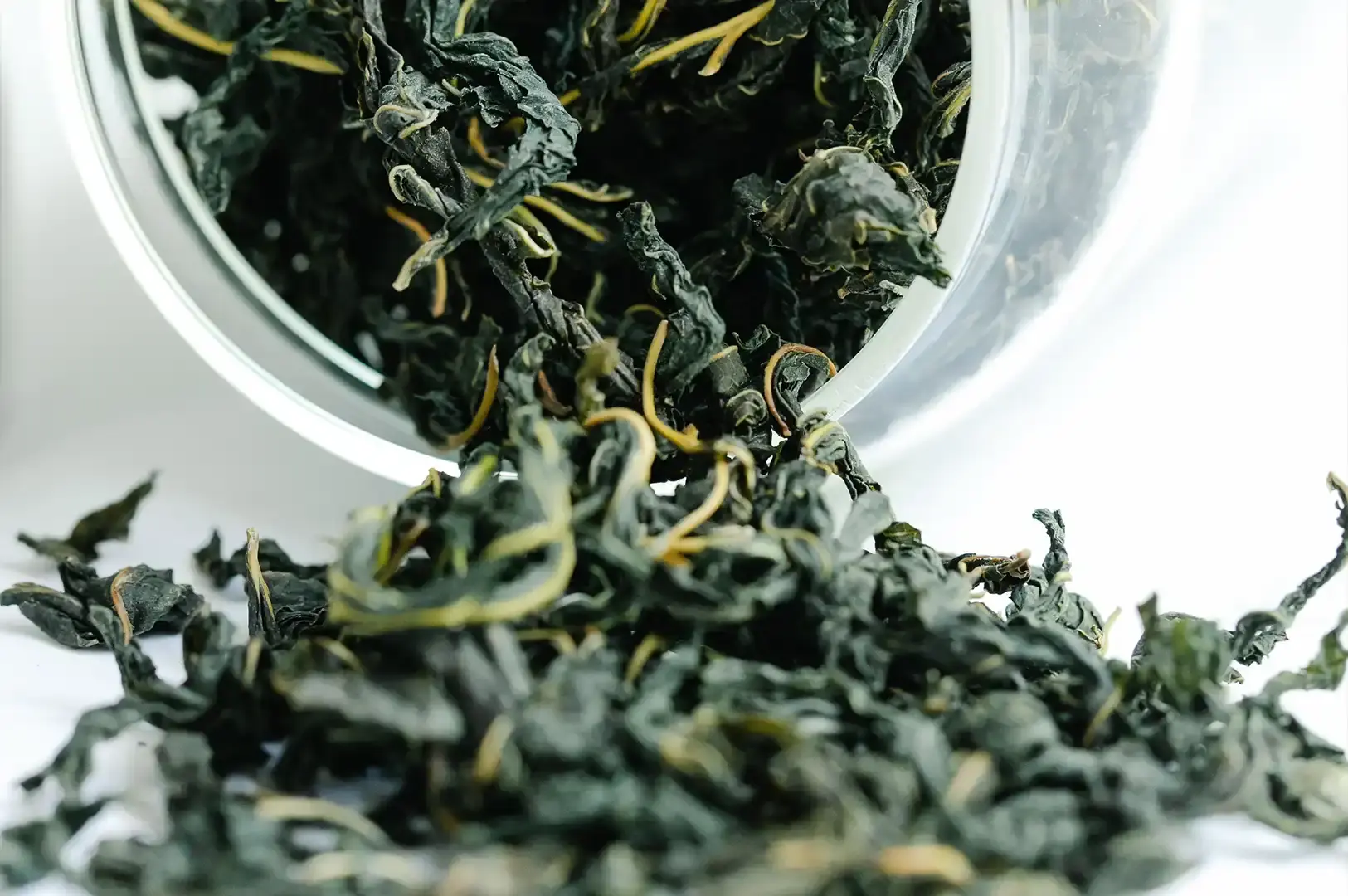 This detailed close-up shot presents dry, dark, and tightly curled Tie Guan Yin tea leaves spilling out of a clear glass container onto a white background. The image focuses on the intricate, rolled structure of the leaves and their variegated colors, providing a clear visual of the tea's raw, pre-brewed state.