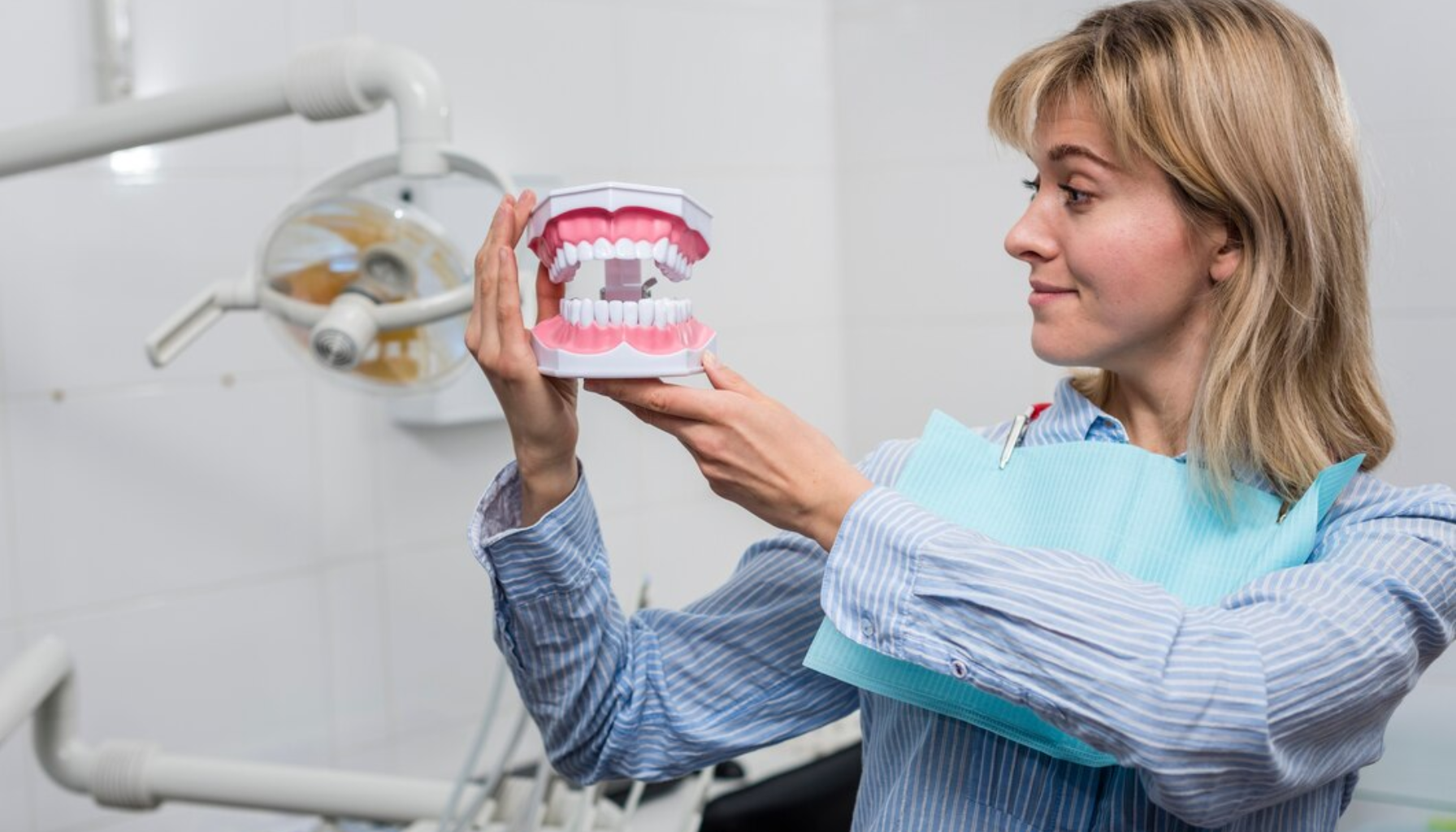 Kvinne i tannlegestol holder en stor tannmodell og ser på den mens hun smiler, omgitt av tannlegeutstyr i klinikk.