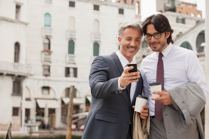 Two businessmen standing outside looking at a cell phone. 