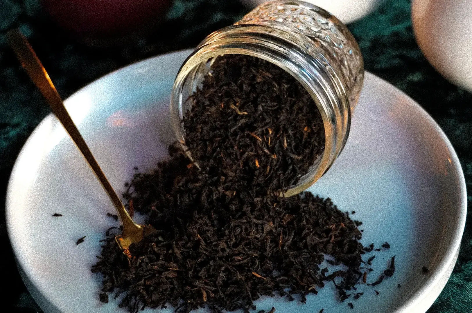 A top-down, slightly angled close-up shows a clear glass jar tipped over on its side upon a flat white ceramic plate, spilling a large pile of loose-leaf black tea onto the surface. The dried tea leaves are dark brown and finely twisted, mixed with a few small, lighter tan twig pieces, while an elegant, slender gold spoon rests on the left side of the plate, partially buried in the scattered leaves.