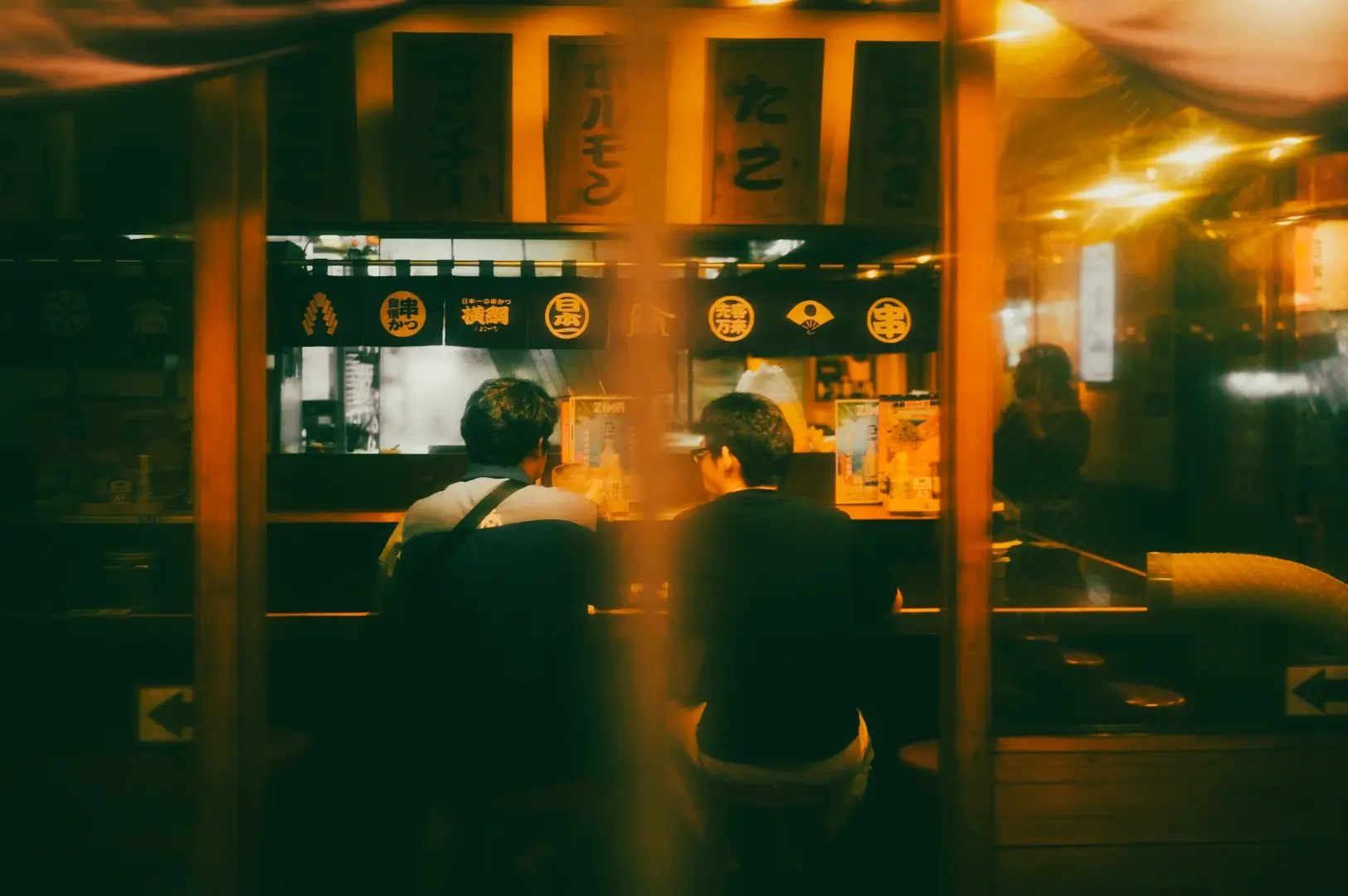 Eye-level exterior-through-window shot of a cozy Japanese izakaya bar showing counter seating with patrons, warm amber lighting, hanging signage with Japanese characters, and an intimate late-night dining atmosphere typical of top izakaya restaurants in Singapore.