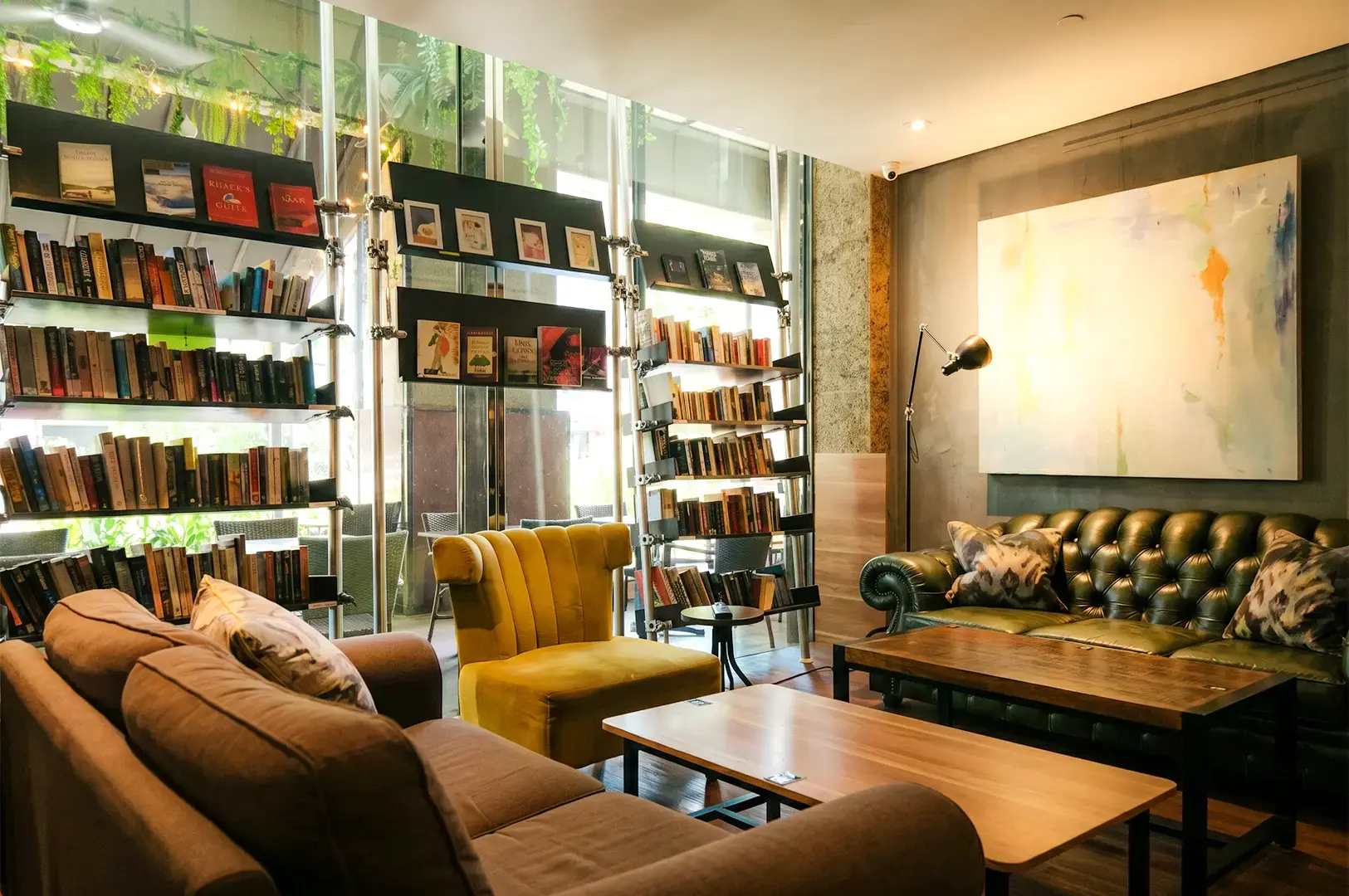 A wide angle shot of Book Cafe's interior, with its book selection arranged on shelves by the glass windows and the comfortable cushioned chairs and wooden tablle in the middle decorated with modern art hanged on the wall.