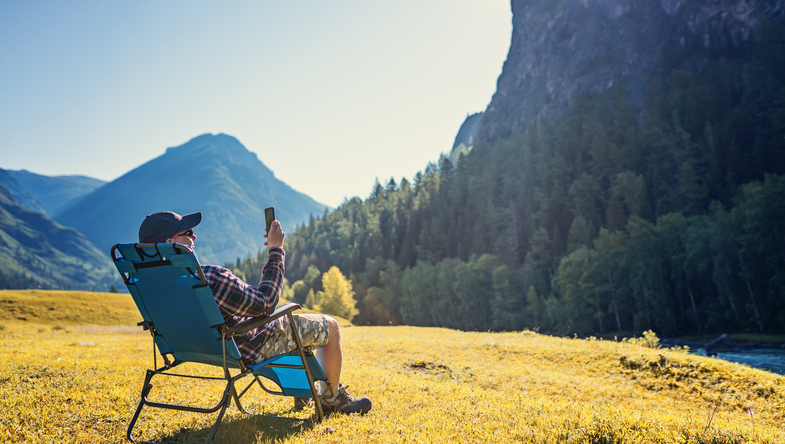 Man sitting in a lawn chair on a mountain looking at his cell phone. 