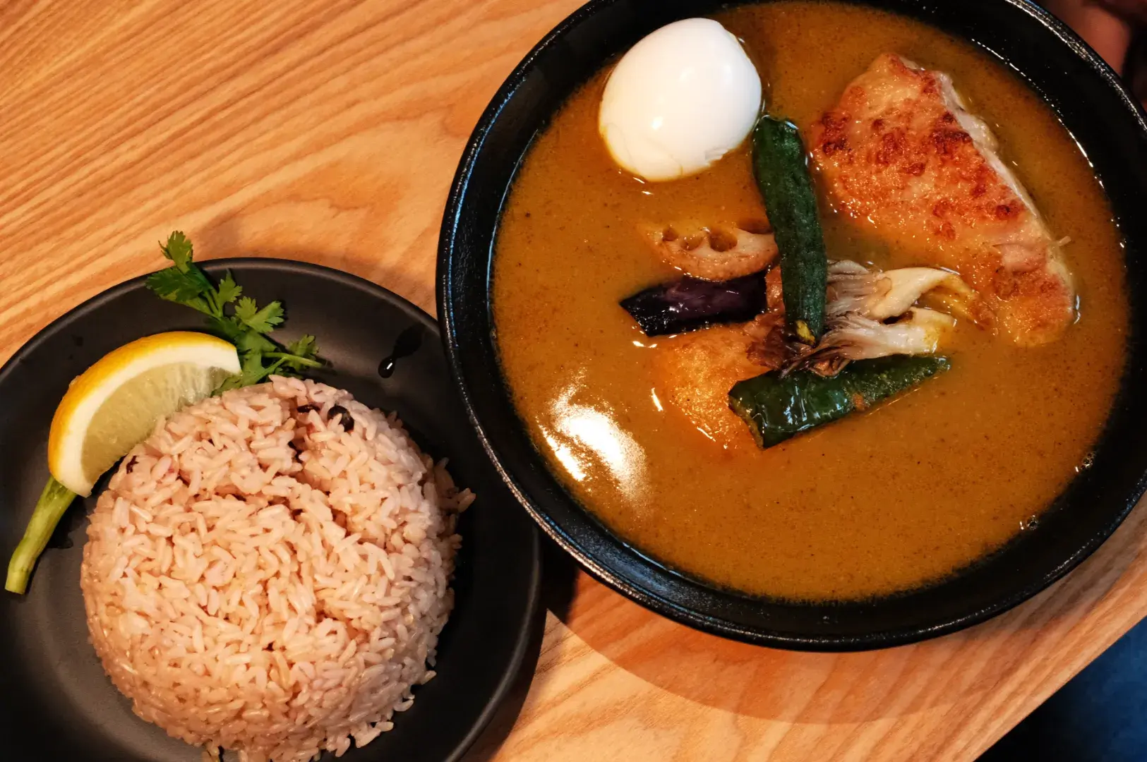 A bowl of curry with a piece of chicken, vegetables, and a soft-boiled egg, accompanied by a plate of brown rice and a lemon wedge.