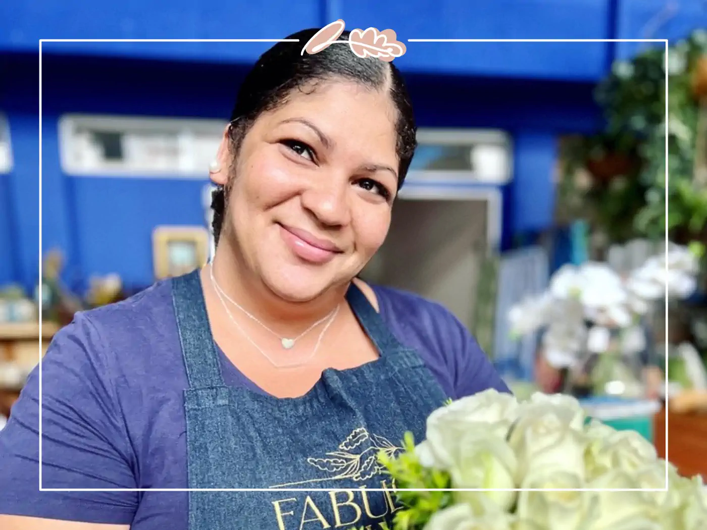 A cheerful woman in a blue apron holding a bouquet of white roses and greenery, standing in a flower shop with a blue background. Fabulous Flowers and Gifts.