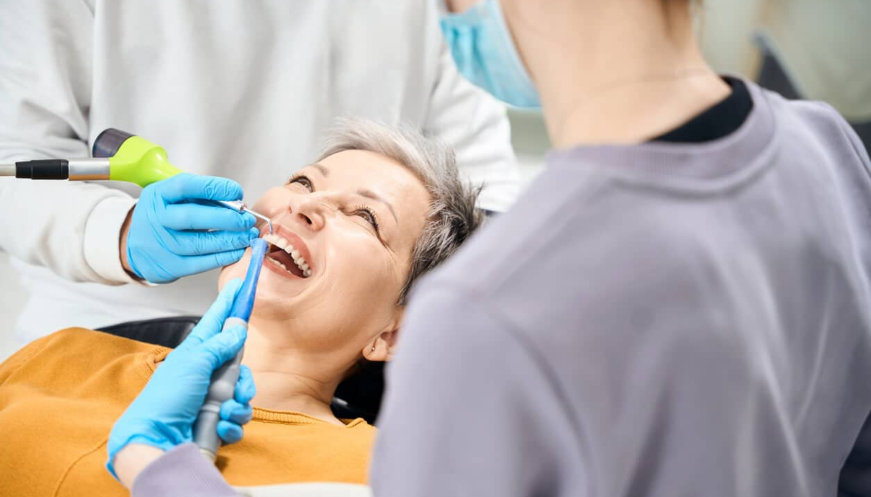 Blemme på Tannkjøttet: Når Små Hevelser Er Et Tegn på Irritasjon eller Infeksjon 18 Tannpleier behandler smilende eldre kvinne i tannlegestol med instrumenter mens kollega assisterer under rutinemessig tannbehandling i moderne klinikk
