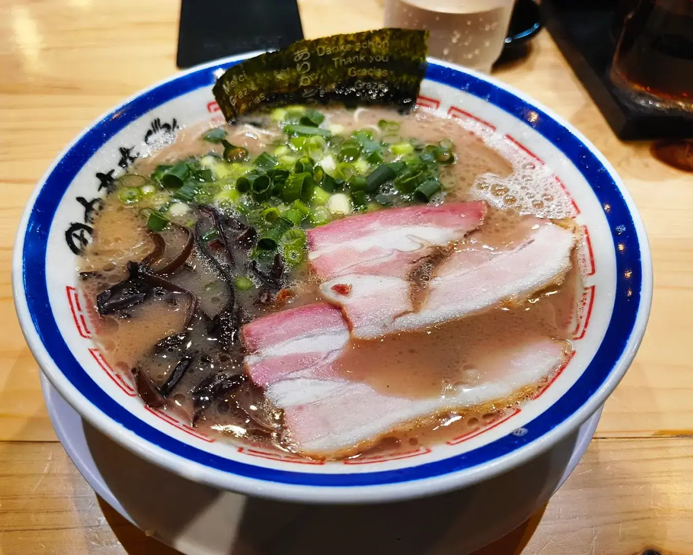 A bowl of ramen with slices of pork, green onions, and seaweed on a wooden table. The broth appears rich and creamy, creating a cozy atmosphere.