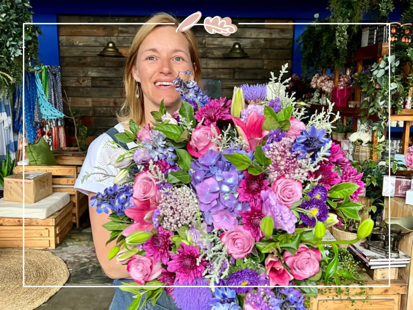 A woman with blonde hair, wearing a white shirt, holding a large, colorful bouquet of pink, purple, and blue flowers, smiling broadly.