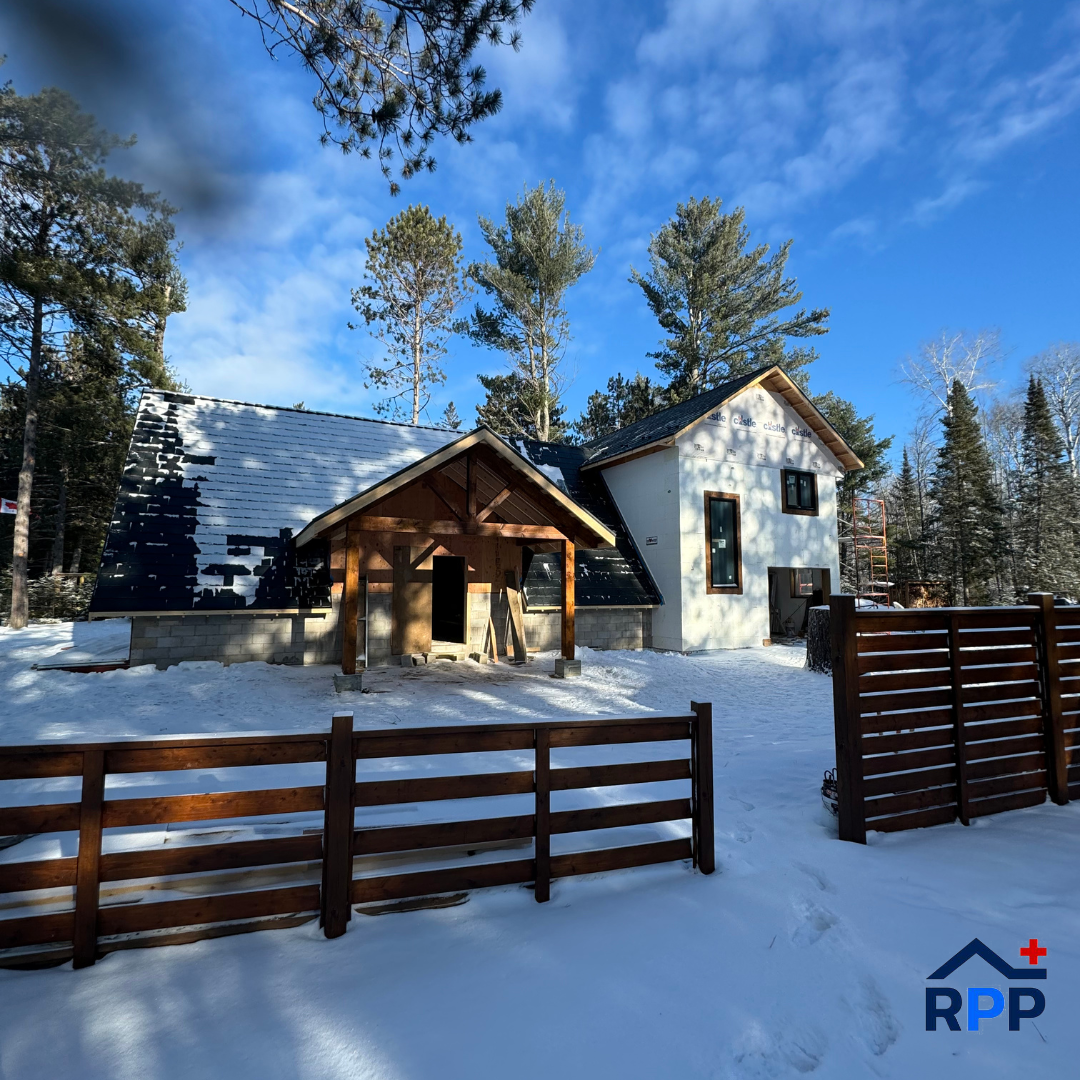 A snowy landscape featuring a modern cabin with a steep metal roof and large windows, surrounded by tall pine trees and a wooden fence under a clear blue sky.