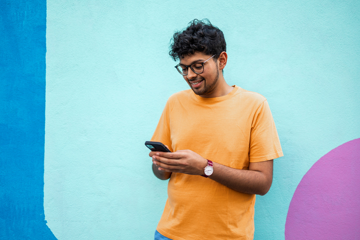 Young man in an orange tee shirt standing in front of blue wall and sending a text.