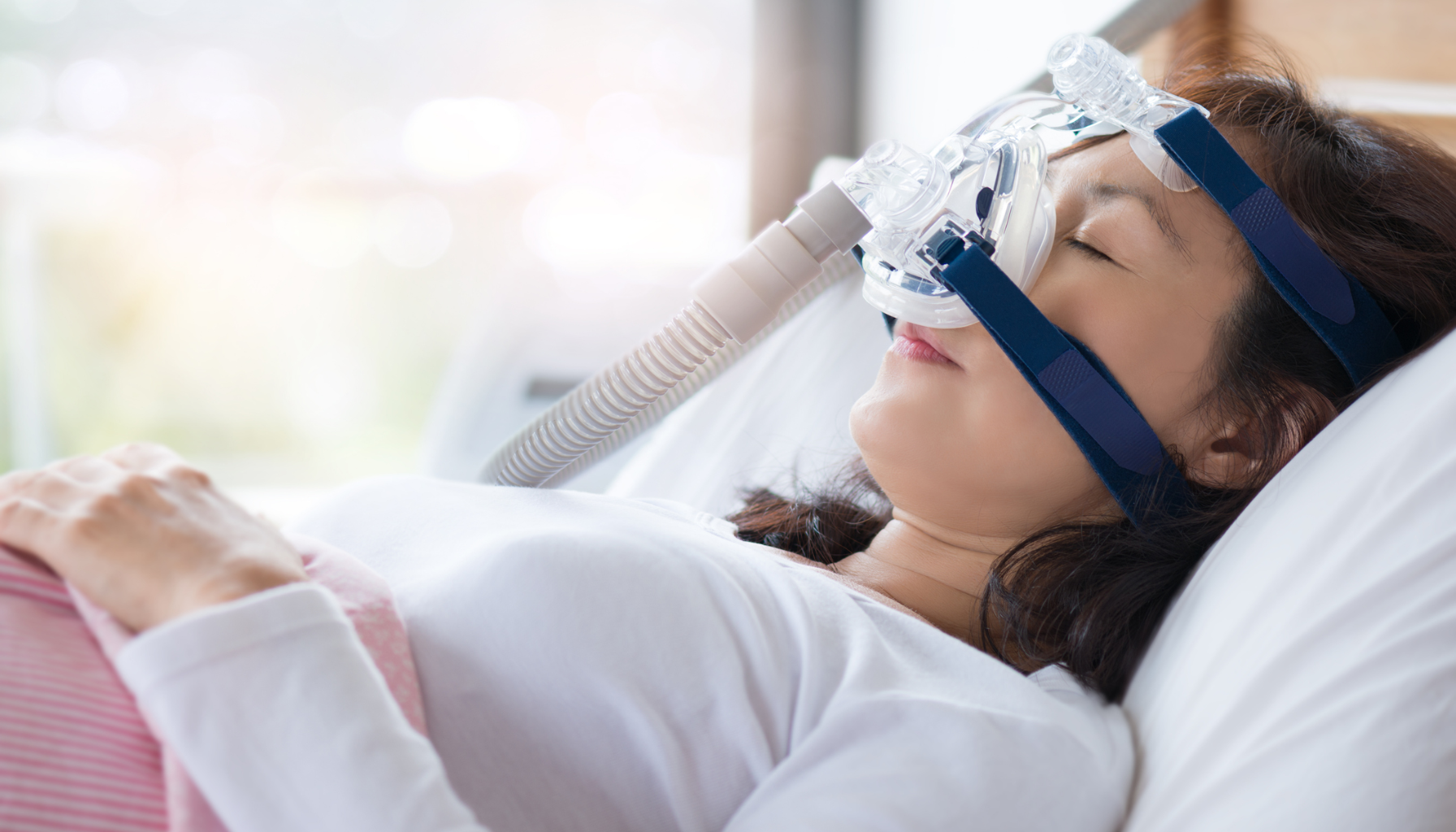 Kvinne ligger i seng med CPAP-maske; slange og maskin synlige ved sengen.
