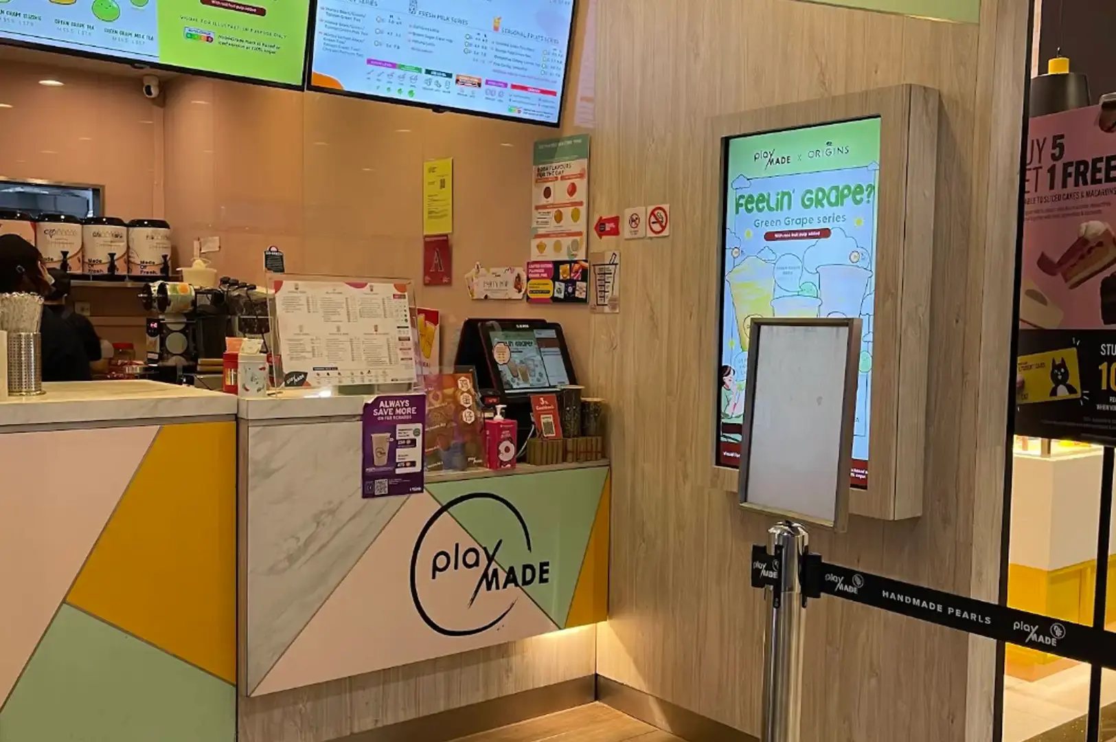 A clean, modern PlayMade bubble tea shop counter accented with light wood paneling, a pastel geometric front desk, digital overhead menu screens, and a vertical digital sign promoting the "Feelin' Grape?" green grape series next to a stanchion queue line.