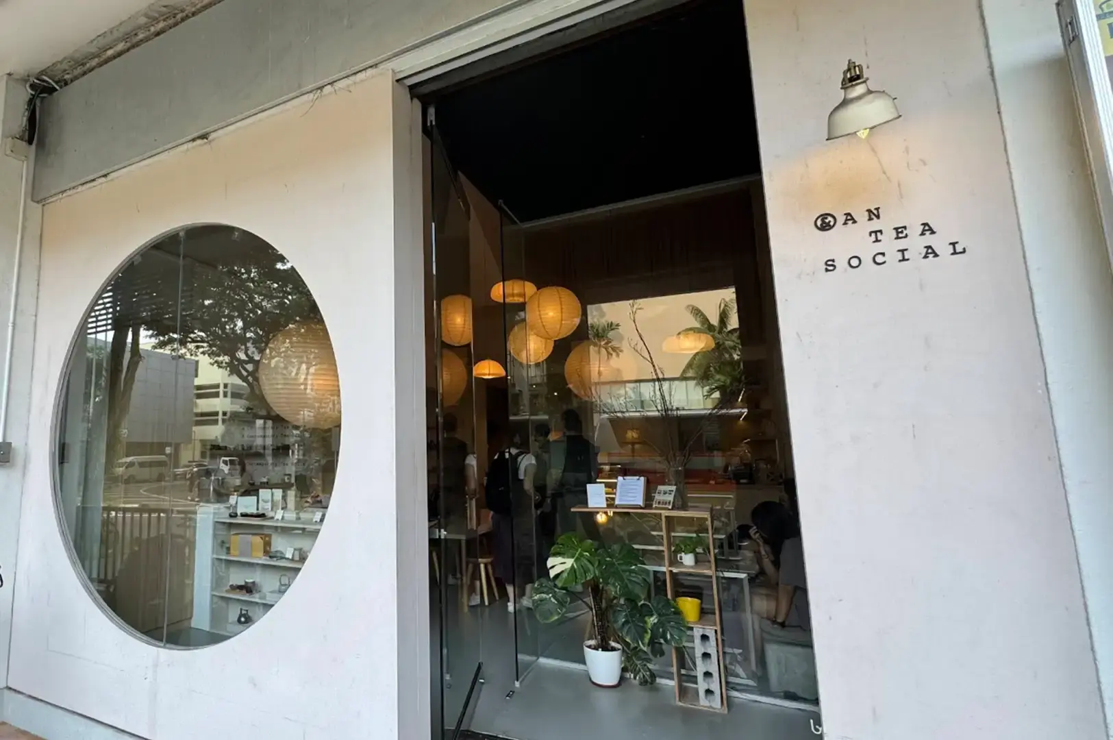 The storefront of Anteasocial features a clean, white minimalist facade with a large circular window on the left revealing warm interior shelves with teaware. On the right, black block lettering spells out "AN TEA SOCIAL" below a small, vintage-style sconce light. The glass entrance door opens into an inviting interior illuminated by warm, spherical paper lantern lights suspended at varying heights, with potted greenery framing the threshold.