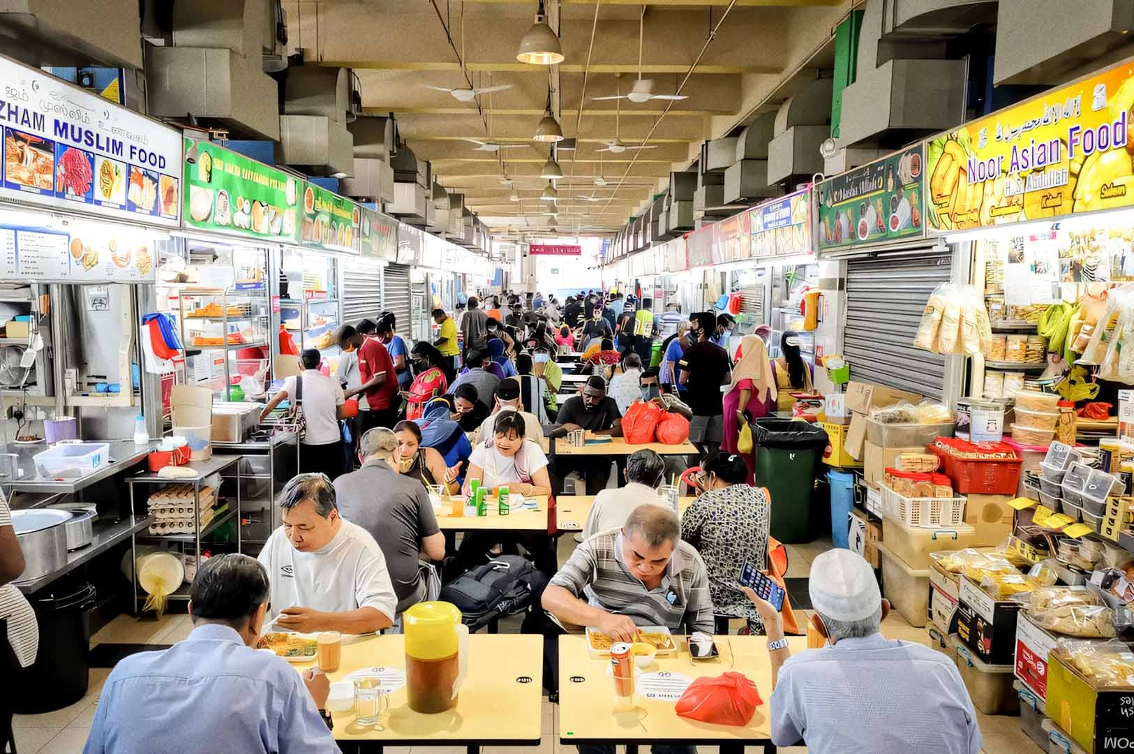 A busy hawker center at Tekka market filled with people eating at shared tables and food stalls lining both sides of the walkway.