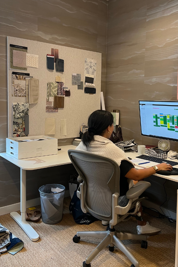 Designer sitting at a desk in a wallpapered office, working on a large monitor beside a material board.