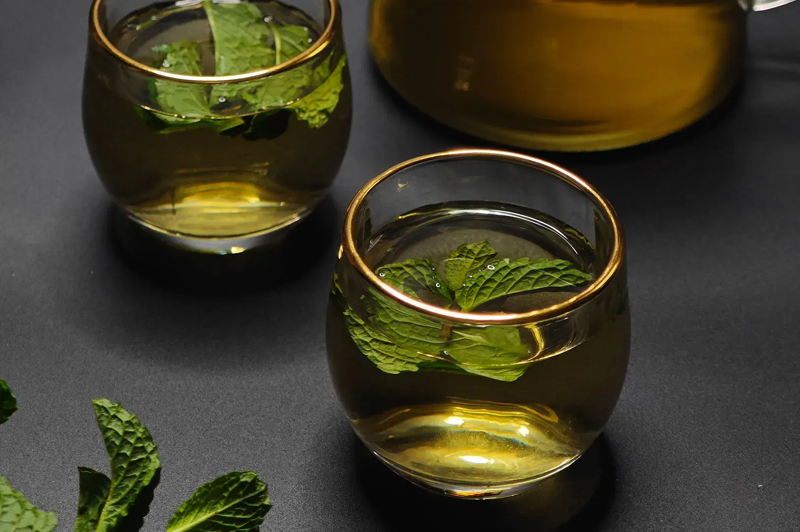 Two gold-rimmed glass cups of fresh mint tea on a dark surface.