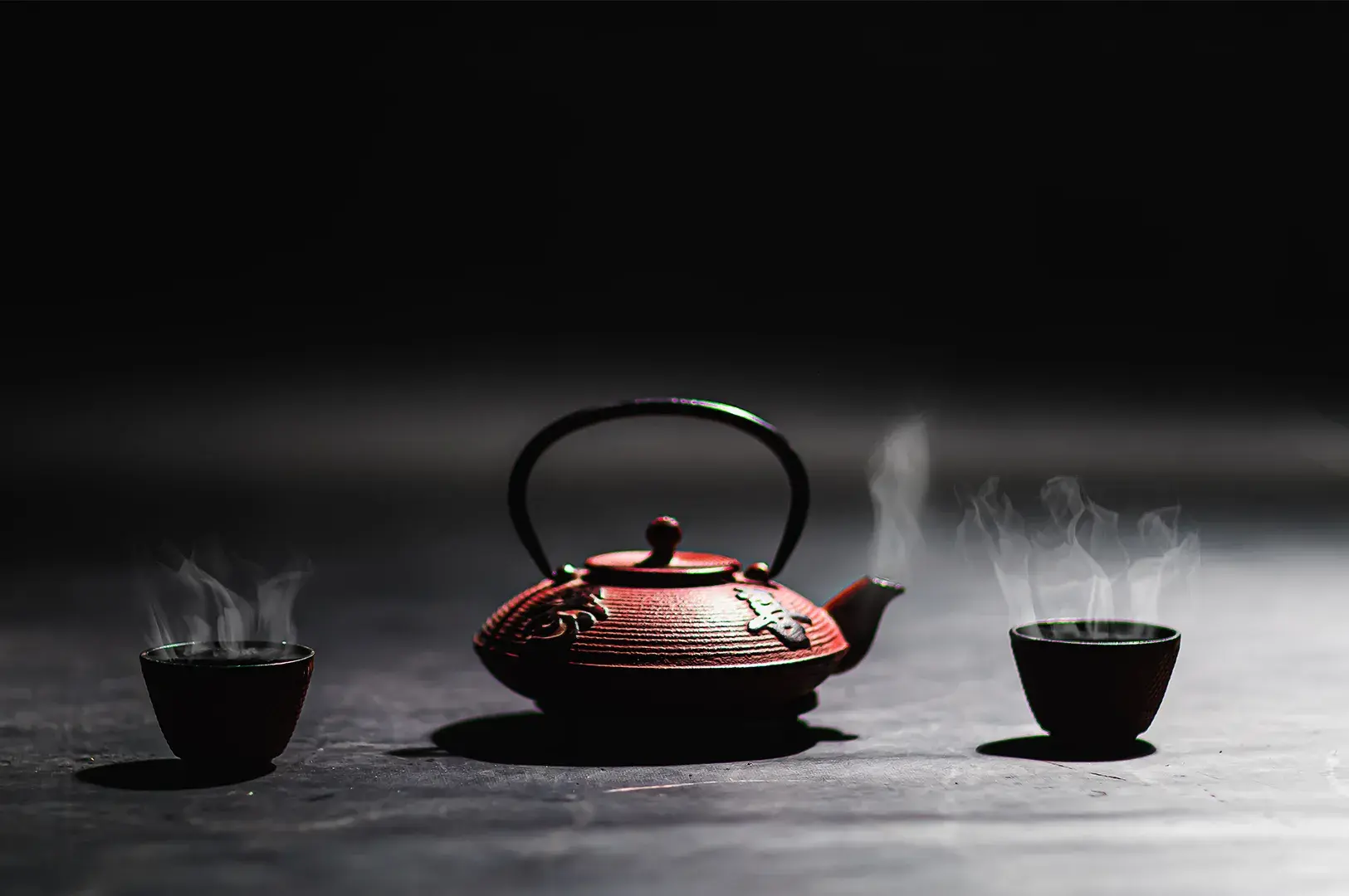 Red cast-iron teapot and steaming cups set against a dark background.