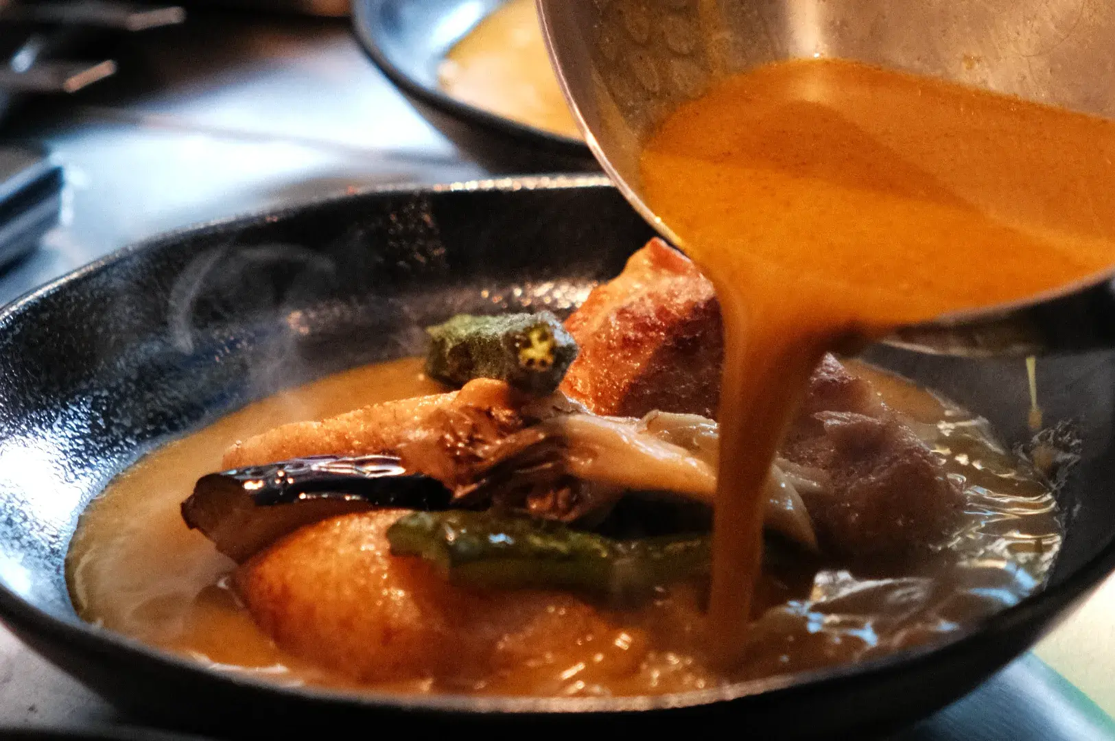 A cook pours rich orange curry sauce into a black pan of simmering chicken and vegetables, filling the kitchen scene with warmth and appetite.