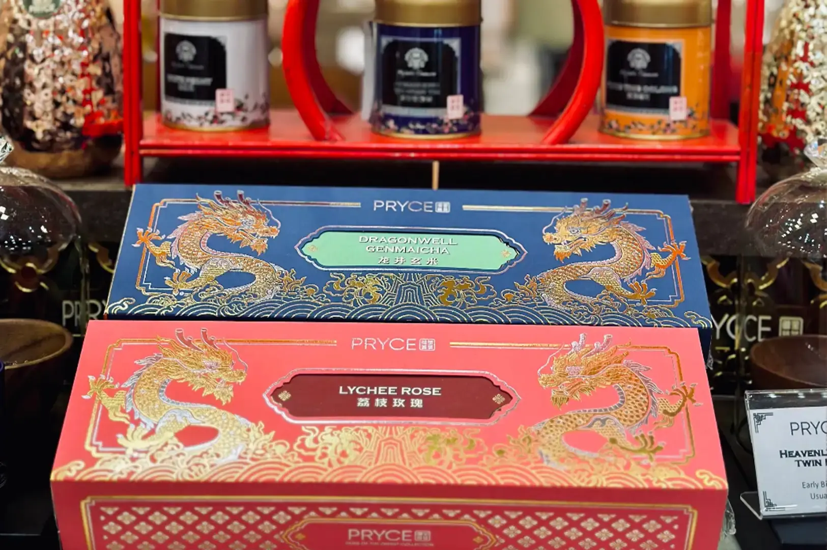 Two long, vibrant tea gift boxes featuring intricate gold dragon illustrations—one in royal blue labeled "DRAGONWELL GENMAICHA" and one in coral pink labeled "LYCHEE ROSE"—are stacked prominently on display. In the background, three tea canisters in white, dark blue, and orange sit on a red wooden display shelf.