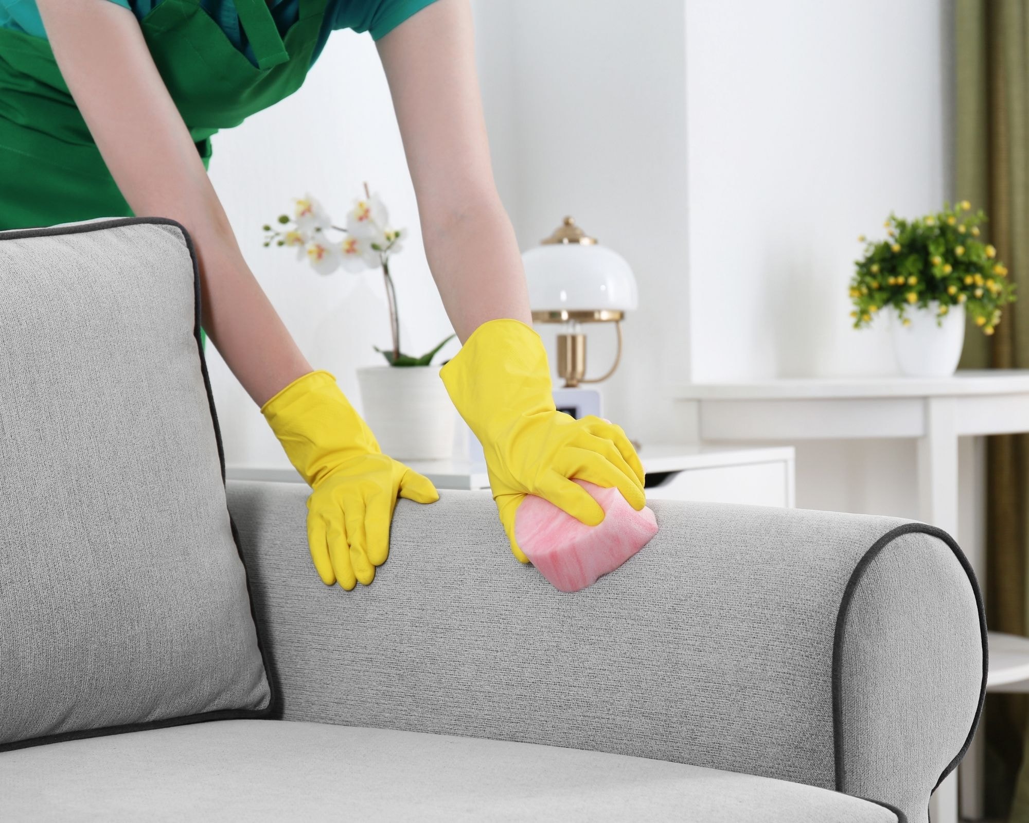 Person wearing yellow rubber gloves cleaning a light gray fabric sofa with a pink sponge in a bright, tidy living room.