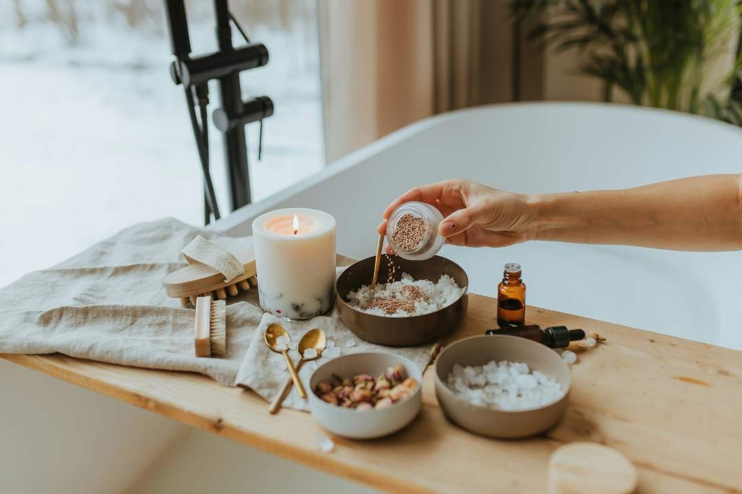 Hands adding salt to a bowl near a candle, essential oils, and dried flowers for a relaxing bath experience.