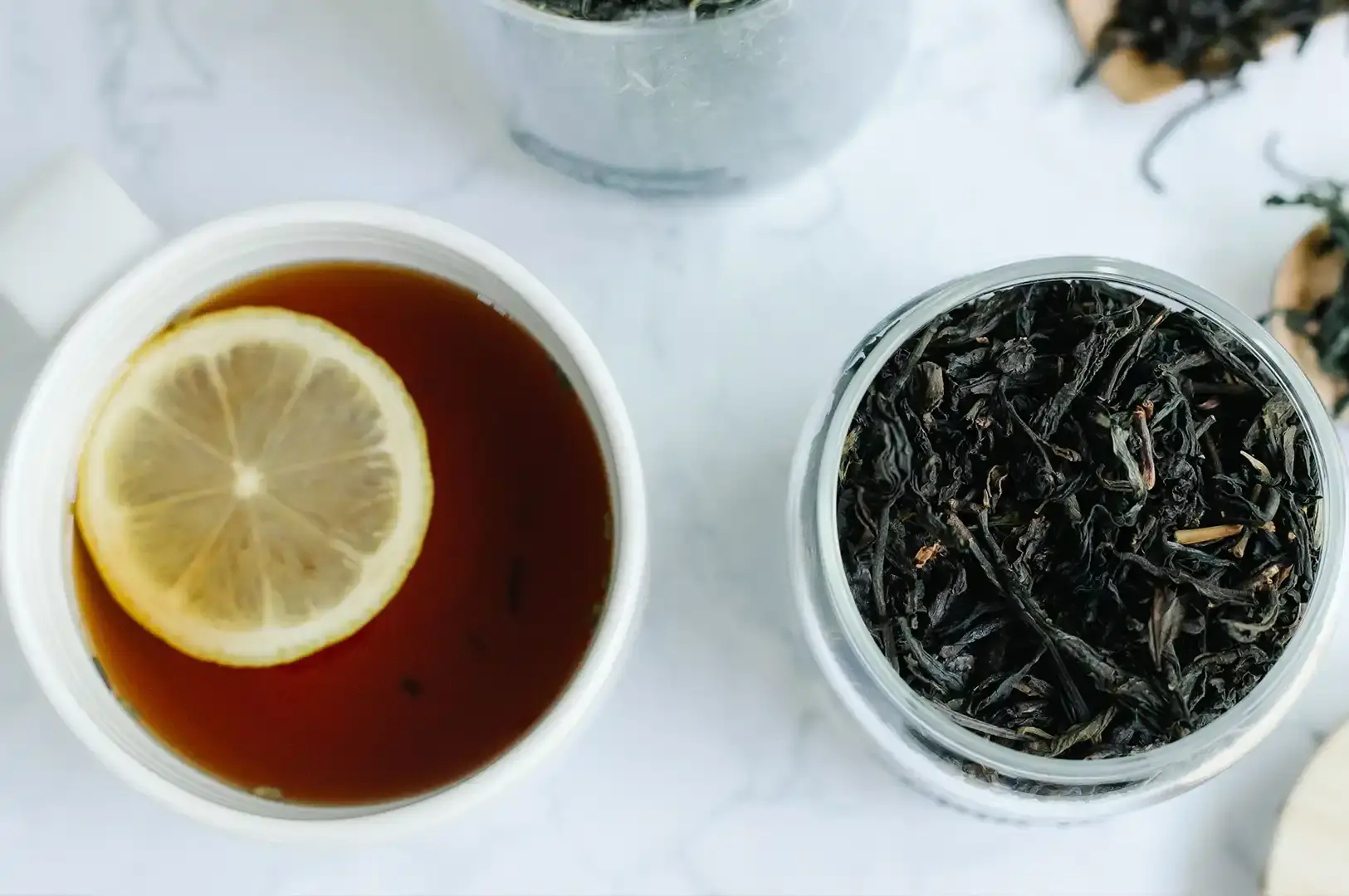 A bright, minimalist scene showing a white cup of amber-colored tea with a slice of lemon floating on top, placed next to a glass jar filled with dark Lapsang Souchong tea leaves, set against a clean, marble-patterned countertop.