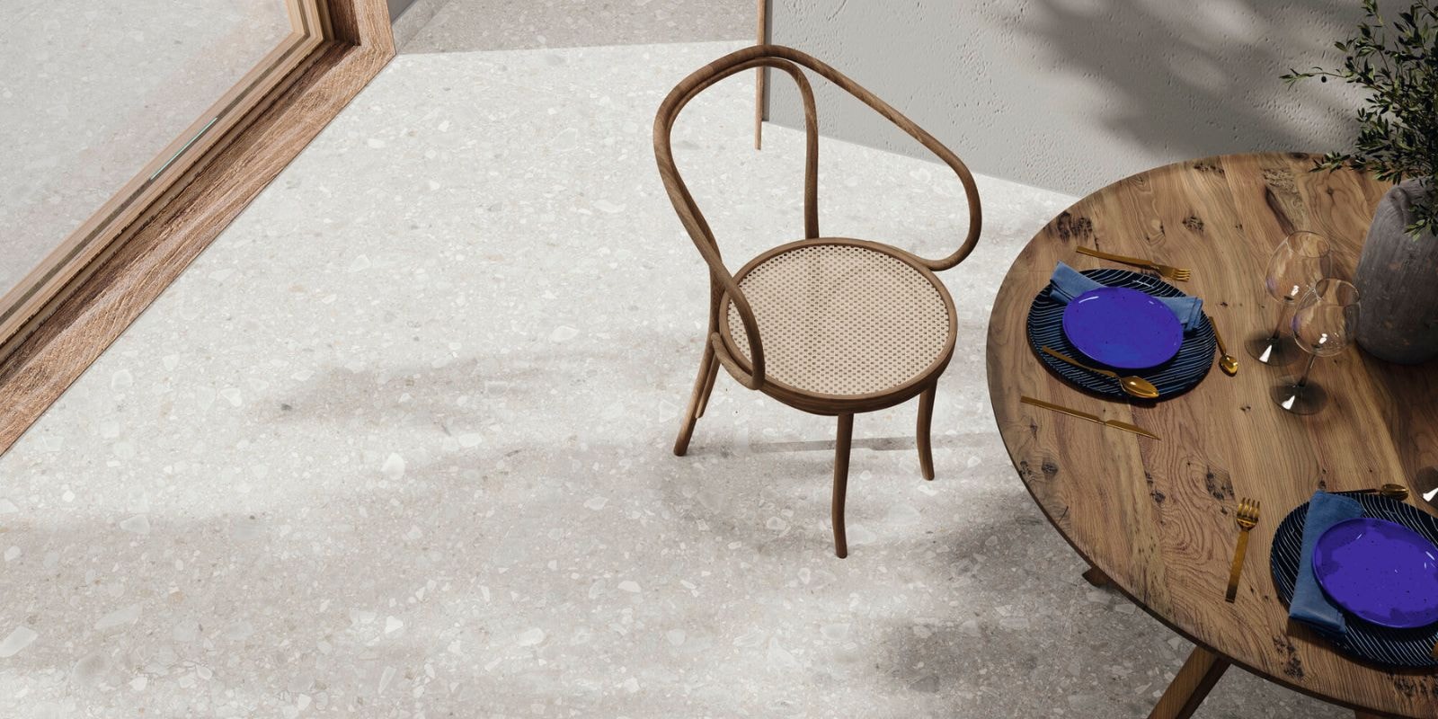 Light terrazzo floor tiles with a speckled pattern of white and beige chips, paired with a wooden chair and a round wooden dining table set with blue plates and gold cutlery.