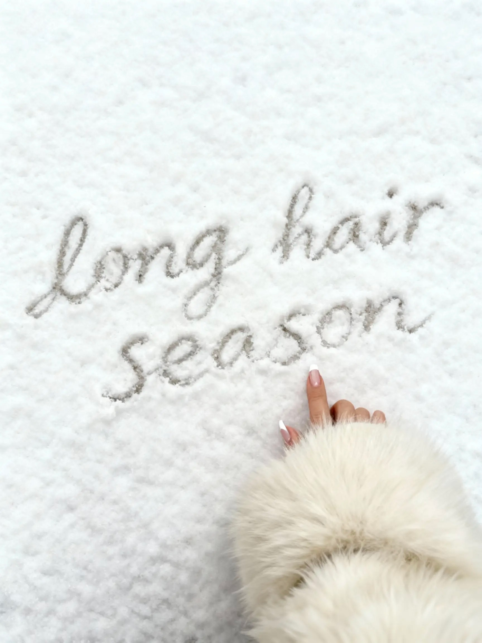 Hand in einer flauschigen Winterjacke schreibt den Schriftzug ‚long hair season‘ in frischen Schnee – winterliches Motiv für Haarpflege, gesundes langes Haar und saisonale Selfcare.