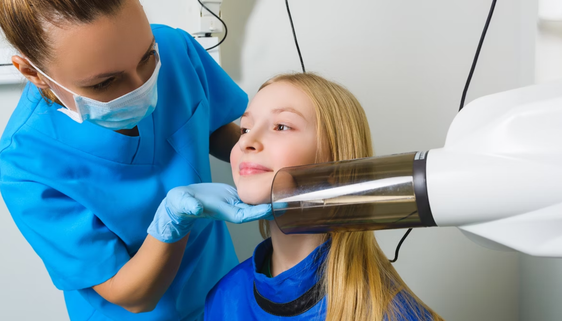 Tannhelsepersonell posisjonerer ung jente for tannrøntgen med moderne utstyr, iført verneutstyr og blå beskyttelsesvest.