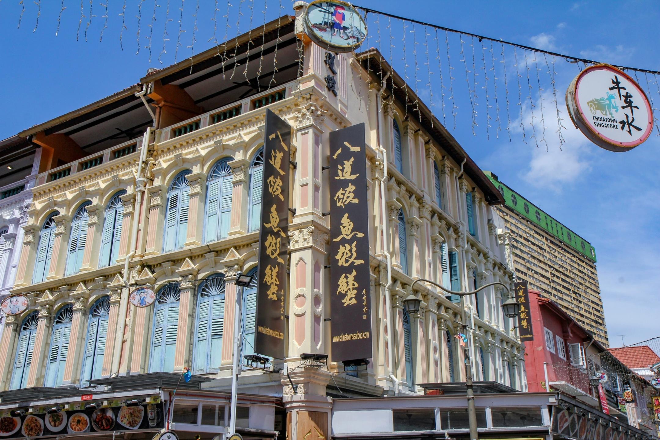 This image captures an ornate corner shophouse characterized by its cream-colored facade, arched blue-shuttered windows, and large vertical black banners bearing golden Chinese calligraphy. Overhead, circular signs and strings of lights hang against a bright blue sky, while a towering yellow residential complex is partially visible in the background, contrasting with the historic architecture.